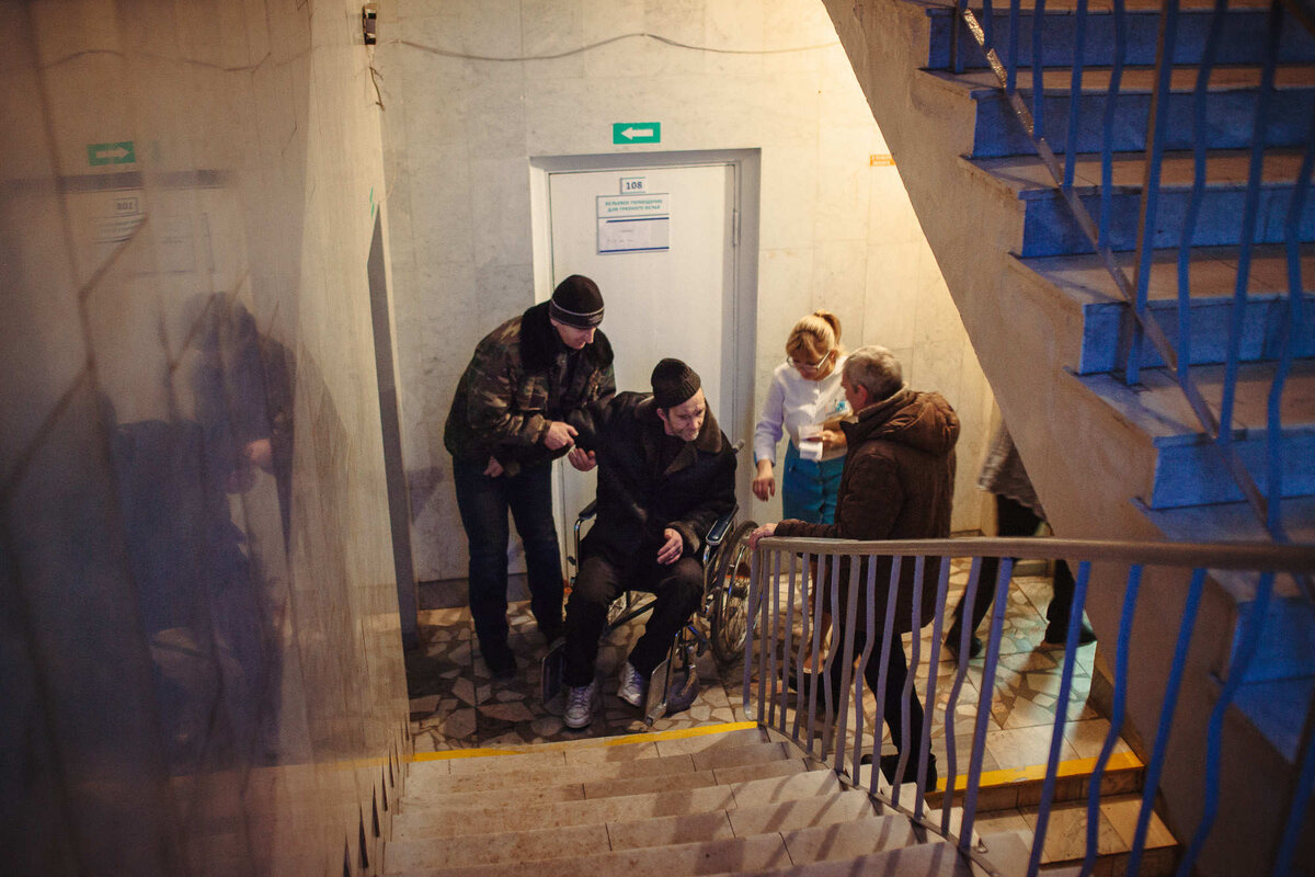 Загурак Игорь (на коляске) пытается попасть к врачу на второй этаж. После черепно-мозговой травмы у Игоря нарушена работа опорно-двигательного аппарата. Без помощи работников поликлиники он не может подняться на нужный этаж.Фото: Александр Васюкович, «Имена»