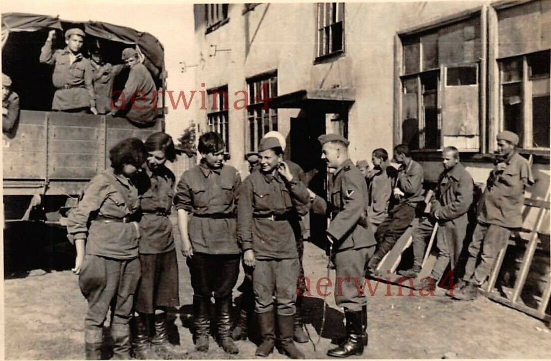  Группа военнопленных женщин у госпитального (?) здания. Справа раненные военнопленные греются на солнце, в бортовой машине военнопленные здоровые. Все с интересом наблюдают за беседой немца с женщинами и еще за какой-то, скрытой от нас сценой на заднем плане.
