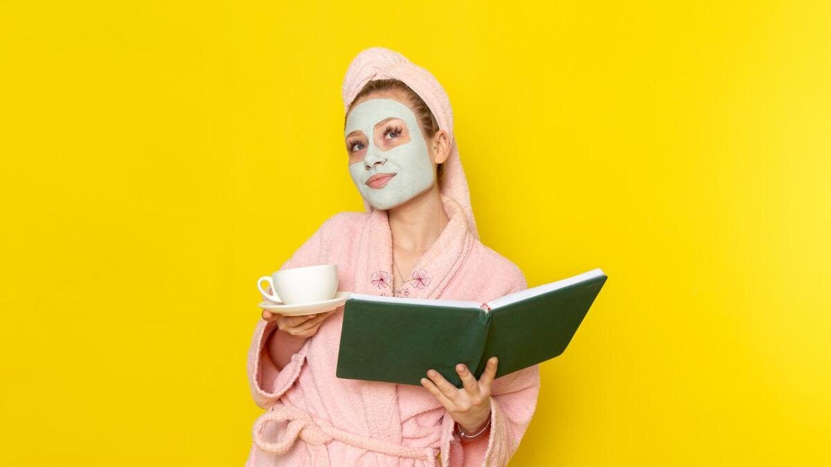    Девушка в халате с белой маской на лице держит книгу и чашку:Freepik/KamranAydinov