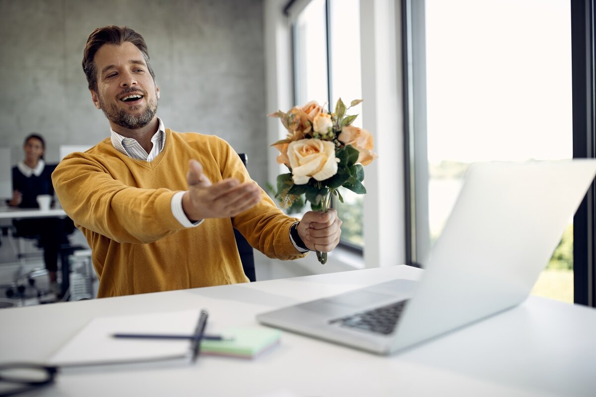    Happy businessman using laptop and making video call while holding bouquet of flowers in the office.