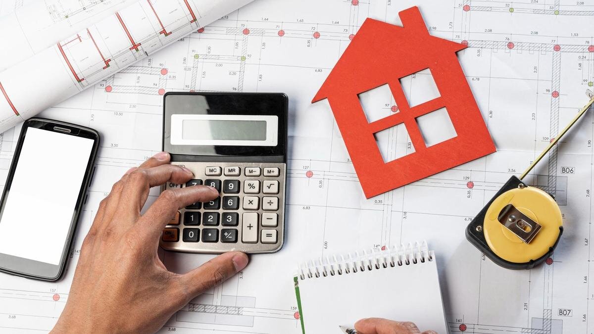   На чертеже лежит телефон, кальулятор, рулетка, блокнот, макет дома и рука:Freepik/Freepik