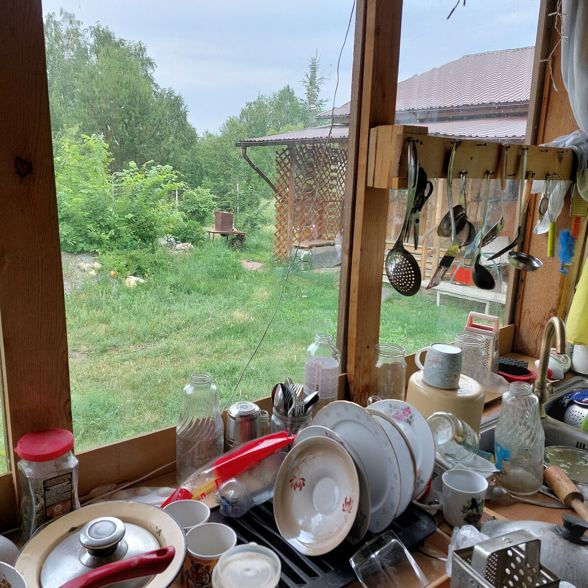 Мы оборудовали себе довольно большую летнюю кухню на веранде. Как-то сил у меня уже меньше стало накрывать на кухне в доме или на открытой террасе: преодолевать крылечки и проч. . Хотя и там, на уличной террасе напротив,  иногда мы с удовольствием садимся за стол, если компания большая. Тем более, что в этом году вообще не получалось полноценно на воздухе спокойно собираться: старший внук сдавал ОГЭ, младшие внуки тоже были заняты. Хотя одну неделю на даче они провели с полным восторгом. А сейчас у нас еще начались комары и мошки.  Так что в то время когда веранда еще строилась (а она и сегодня в процессе...), то становилось ясно, что это место у нас получается с отличным обзором территории и очень удобное для самой разной коммуникации. Поэтому я и попросила мужа сделать на веранде дачно-кухонныйй угол удобным именно для себя, потому что мне чаще всего приходится что-то готовить и , одновременно, быть в курсе дел младших внуков. Вот даже поварешки мы повесили так, чтобы сразу можно  было быстро посушить всё после мойки. И мойки у нас две рядом. Одна на веранде и другая через стенку у крыльца (зимой они отключаются), чтобы первую грязь и землю можно было оставить за пределами кухни. В доме  у нас тоже есть кухня, но летом там совершенно не хочется собираться... Лето - это ведь такая вольница в кухонных делах, что возможны разные варианты. Тем более, что и барбекюшница у меня оказывается в поле зрения. А она, кроме своей основной функции - шашлыков - служит еще и домиком для огромного количества машин и игрушек внуков. И им удобно, и нам... Чтобы им сразу выехать оттуда с помпой и потом быстро туда же заехать, если это нужно: дождик, домой уезжают...