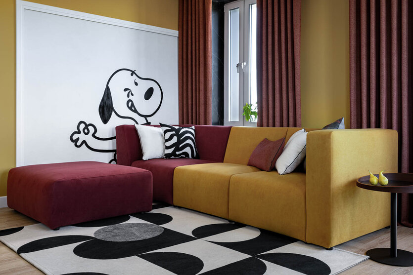 Визуальная доминанта гостиной — чёрно-белая роспись со Снупи, выполненная по эскизам автора проекта. Диван и пуф также выполнены по эскизам дизайнеров мастерской «Rubini». Ковер из хлопка и полиэстера, Carpet Decor.