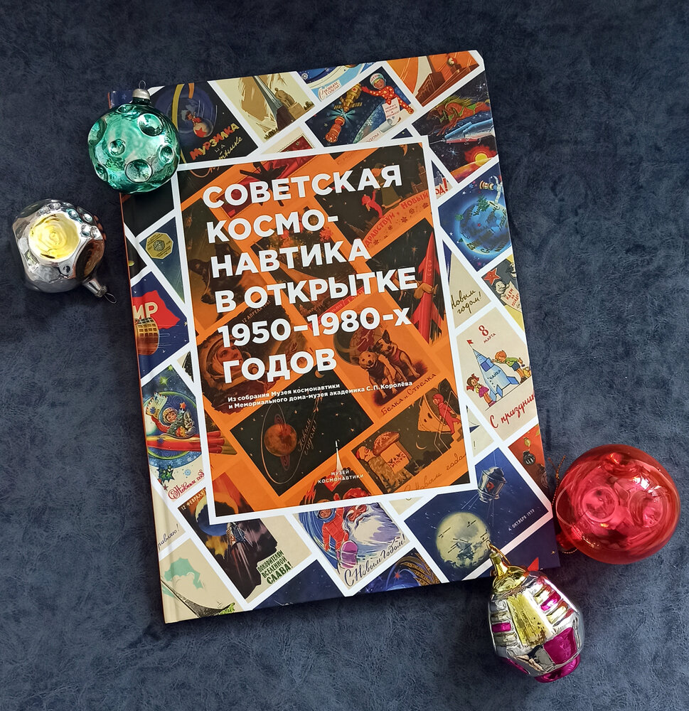 Фото автора. Игрушки из коллекции автора на фоне книги Советская космонавтика в открытке 1950-1980-х годов. Из собрания Музея Космонавтики и Мемориального дома-музея академика С.П. Королева
