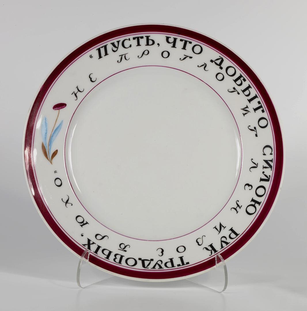 Тарелка с надписью: "Пусть, что добыто силою рук трудовых, не поглотит ленивое брюхо".  1918. Видим тарелку с примитивным декорированием (пурпуровой каёмочкой) и лозунгом по кругу. Из дополнительного декора - осторожный маленький стилизованный цветочек. Начало пути. Роспись надглазурная полихромная. Размеры: 2,9*23,9 см. Собрание Эрмитажа. Предмет поступил в 2002-м году. Передан в составе коллекции Музея Фарфорового завода. Инв. номер Мз-С-3149. Изображение взято из каталога Эрмитажа. Листайте галерею вправо.