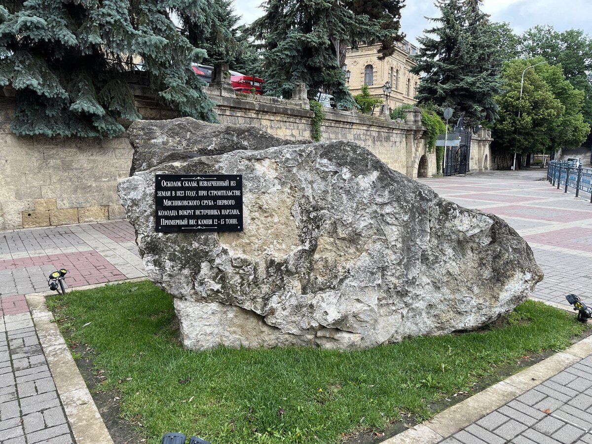 Надпись гласит: "Осколок скалы, извлечённый из земли в 1823г, при строительстве Мясниковского сруба, первого колодца вокруг источника нарзана. Примерный вес камня 12-15 тонн". 