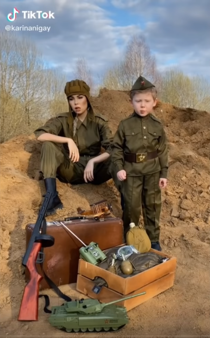 Яркий пример победобесия в лице мамочки, сидящей в привычное ей манере, ведь чем торгует, тем и поздравляет. Жаль, что ППШ игрушечный...