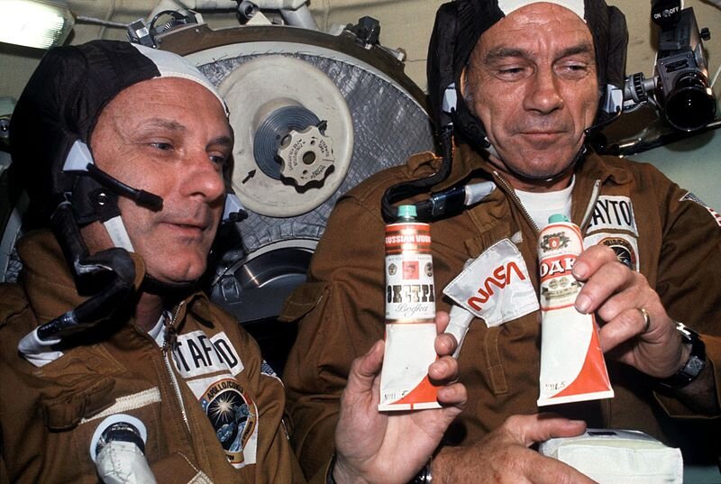 NASA. Public domain. Водка в тюбиках на "Союзе". Astronauts Thomas P. Stafford (left) and Donald K. "Deke" Slayton hold containers of Soviet space food in the Soyuz Orbital Module during the joint U.S.-USSR Apollo-Soyuz Test Project docking in Earth orbit mission. The containers hold borscht (beet soup) over which vodka labels have been pasted. This was the crews' way of toasting each other.