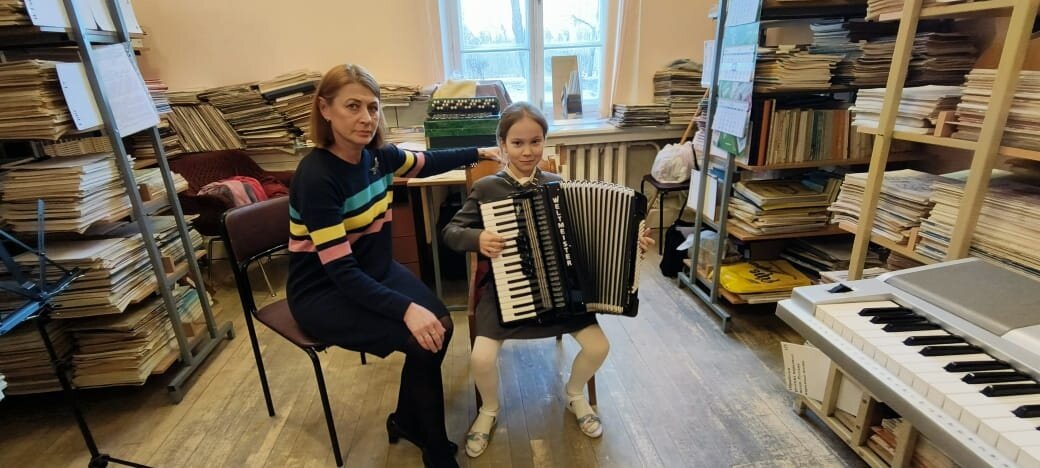 Марина Егоровна занимается с Алёной Сидоренок (моей сестрой) в библиотеке ДМШ №25. За ними стоит стол с аккордеонами и баяном.