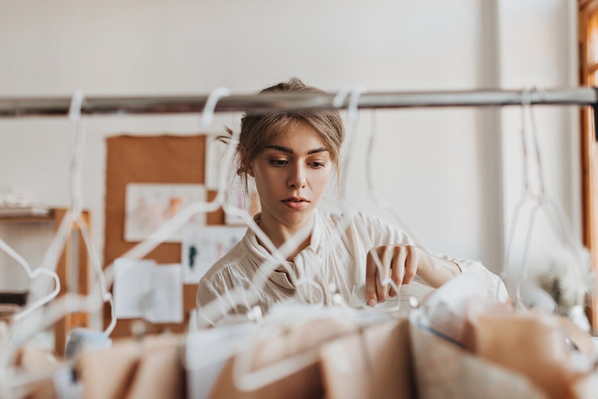    Cheerful woman looks at her clothing samples. Attractive girl in beige blouse works on new fashion collection in white office