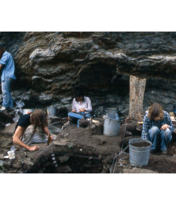 Два археолога на переднем плане копают (тот, что слева, использует ручную метелку). Сидящий археолог настраивает фотоаппарат. Стоящий слева собирается с помощью картографической сетки рисовать карту дна квадрата.