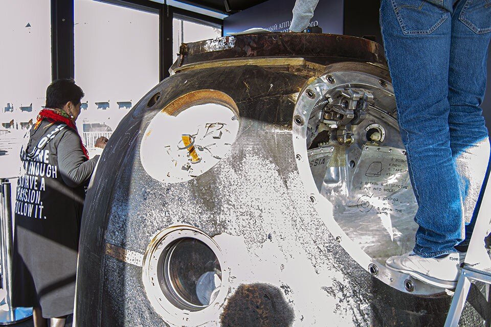     Спускаемый аппарат «Союза МС-18», работники протирают экспонат от пыли (не космической). Яркое апрельское солнце освещает абляционную теплозащиту корабля. После того, как она прошла атмосферу, она сильно обгорела (это нормально). Источник: Роман Белоусов