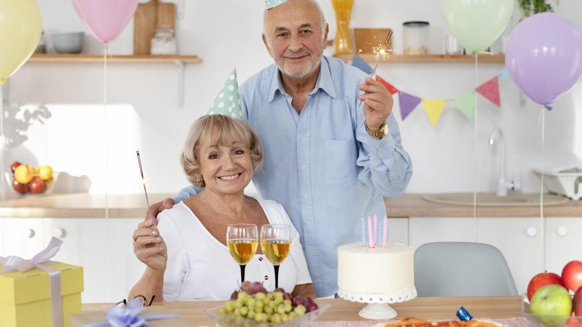   Женщина и мужчина в именинных колпачках улыбаются и держат в руках горящие бенгальские свечи. Женщина сидит за праздничным столом, а мужчина стоит рядом:Freepik