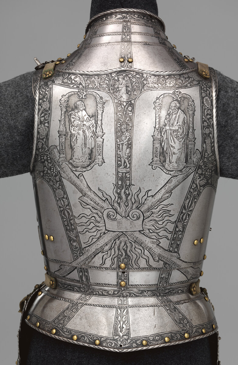 Задняя сторона доспеха украшена скрещенными посохами и огненными клинками, знаками отличия Ордена Золотого Руна, элитного рыцарского общества, членом которого был Фердинанд