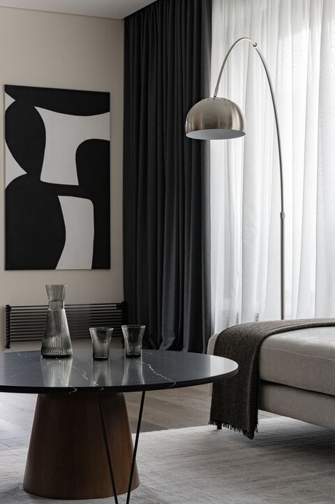 Журнальный столик изготовлен по эскизам дизайнеров, подстолье выполнено из массива дерева и стальной трубки, столешница — из натурального камня. Картина на стене служит акцентным пятном и разбавляет строгую геометрию штор и радиаторов отопления.