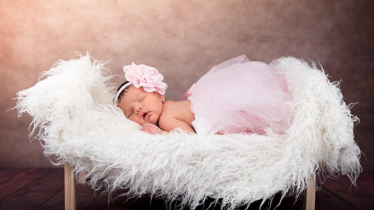    Новорожденная девочка лежит на кроватке с пушистым белым покрывалом. На ней одета розовая пачка и обруч с цветком на голове:Pixabay