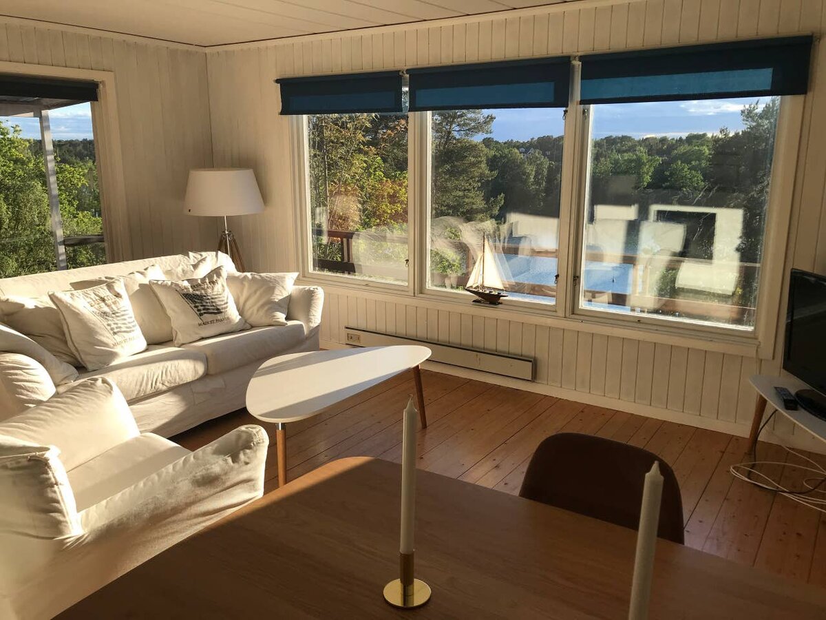 Гостинная. Большие окна по всему периметру комнаты. Стены и потолок окрашены белой полупрозрачной краской, пол - лак.  Белый диван с множеством подушек.  Столик, роллерные шторы и торшер из Икея. Минимализм и практичность во всей красе.