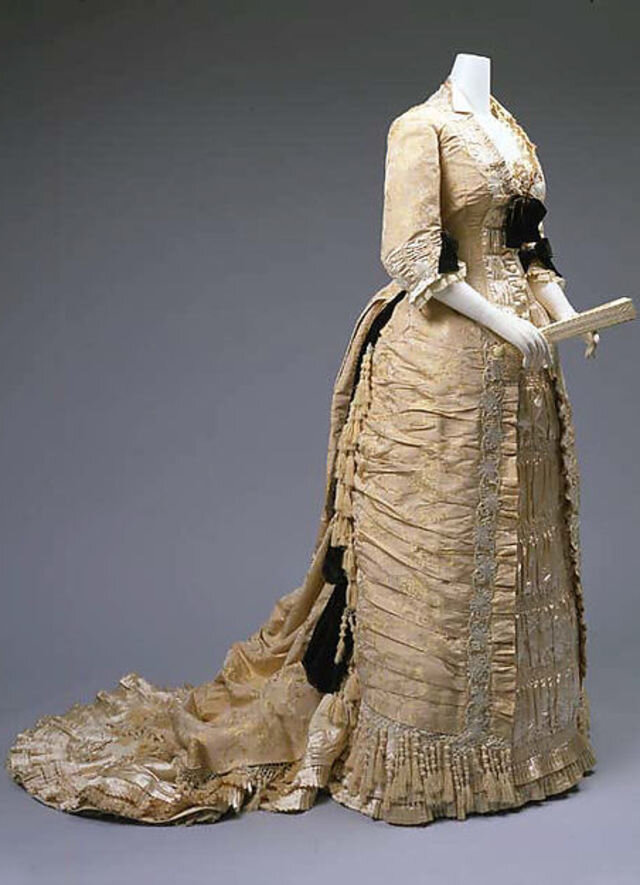 Платье для вечернего приёма или поездки в оперу - но не для бала - середина 1870-х годов. (Время действия романа - 1874 - 1876 гг) В 1870-х и 1880-х годах весь объем юбки собирается сзади. Чтобы удерживать эту форму, под платьем носят специальные конструкции из проволоки. Форма эта называется турнюр, и, по факту за полтора десятилетия сменилось несколько основных форм турнюров, хотя принцип (акцент - сзади) оставался прежним. 